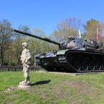 Military tank display with soldier statue in a grassy, outdoor park setting.
