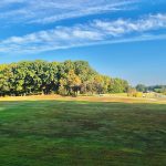 Expansive green golf course with a lush, tree-lined horizon under a bright blue sky.