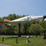 White and orange jet aircraft on display outdoors, set against a backdrop of green trees.