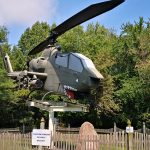 Military attack helicopter displayed outdoors amidst lush green trees and a wooden fence.