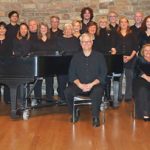 Group of musicians and choir members posing with a grand piano in a warm, rustic setting.