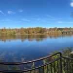 Blue lake reflecting autumn trees under a bright, clear sky with a white fence.