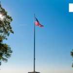 American flag flying on a tall pole against a bright blue sky, framed by green trees.