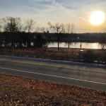 Sunset over a calm lake with trees lining the shore and a road in the foreground.