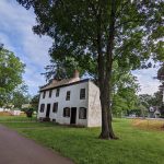 Historic white farmhouse nestled among large green trees on a grassy park path.