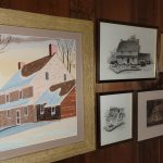 Framed artwork depicting a quaint house scene and various architectural sketches on wood paneling.