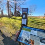 Informational sign near a park bench with trees and open field under blue sky.