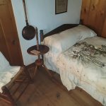 Rustic bedroom with wooden walls, a bed covered in patterned linen, and an antique kettle on a stand.