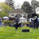 Musician performs outdoors near a white gazebo with greenery and trees in the background.