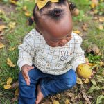 Baby with yellow bow sits on grass, holding a small yellow fruit.