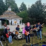 Live band performs outdoors near a gazebo under trees during a daytime event.