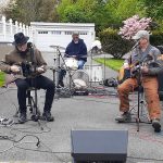 Musicians perform outdoors on a paved driveway with a white garage in the background.