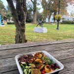 Meal in foil tray on wooden table with park and statue background.