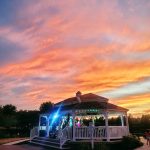 Sunset over a white gazebo with people gathered, vibrant orange and pink sky.