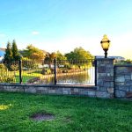 Stone wall and wrought iron gate overlooking a pond on a sunny day.