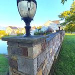 Stone retaining wall with a decorative lantern overlooking a body of water and houses.
