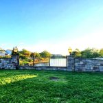 Stone wall and black iron gate overlooking a pond with suburban houses under a blue sky.