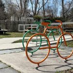 Orange and green bicycle art sculpture on a paved outdoor area with trees in the background.