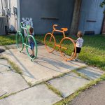Two young children play with colorful, oversized bicycle toys outdoors on a concrete path.