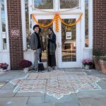 Two people stand outside a brick building with colorful decorations and a patterned welcome mat.