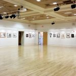 Bright, empty gallery space with light wood floors and framed artwork on white walls.
