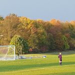 Soccer player on a green field with a goal net, autumn trees in the background.