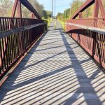 Wooden pedestrian bridge with long shadows stretching across the walkway under a clear sky.