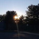 Sunset silhouette over a basketball court with trees framing the bright sun.
