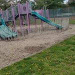 Colorful playground equipment set in a grassy park area with trees and fencing.