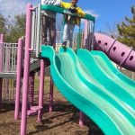 Two boys on a colorful playground structure with a bright blue and green slide under a clear sky.