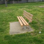 Wooden park bench on a gravel pad in a grassy outdoor area with trees in the background.
