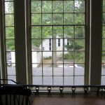 Large window view of a white house and green trees on a sunny day.
