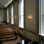 Interior view of a church aisle with wooden pews and tall, arched windows.