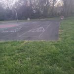 Outdoor basketball court with painted lines, surrounded by grass and trees under an overcast sky.