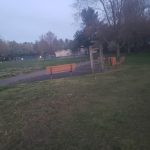 Park scene with grassy field, path, wooden benches, and trees under a cloudy sky.
