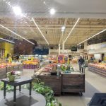 Indoor grocery store aisle with fresh produce displays and bright overhead lighting.