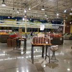 Grocery store interior with produce displays, aisles, and shoppers under bright lighting.
