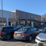 Commercial strip mall with businesses under a clear blue sky, featuring parked cars in the foreground.