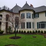 Large, elegant light-colored house with dark roof and mature tree in front.