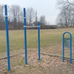 Outdoor fitness equipment set with blue poles and a workout sign in a grassy park area.