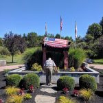 Man stands before a memorial with American flag under clear blue sky.