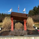 Bronze monument with flags set against a backdrop of trees and dry grasses under a blue sky.
