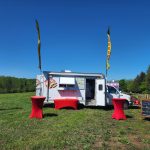 Food truck parked in a grassy field under a clear blue sky with promotional flags.