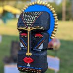 Black tribal mask with gold accents and blue trim, displayed outdoors.