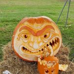 Giant carved pumpkin with a scary face sits on hay in a grassy outdoor setting.