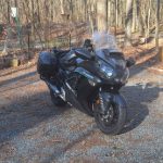 Black sport motorcycle parked on a gravel path surrounded by bare trees in a wooded area.