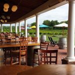 Outdoor patio dining area with wooden tables and chairs overlooking a vineyard under an overcast sky.