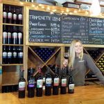 Woman smiles behind bar stocked with wine bottles and a chalkboard menu.