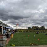 Overcast sky above a grassy field with people gathered near a building and American flag.
