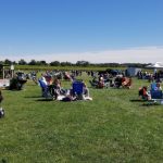 People gather on a sunny green field for an outdoor event with tents and picnic setups.